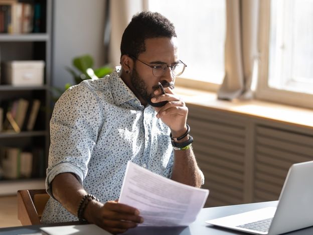 Focused african businessman holding documents looking at laptop doing research