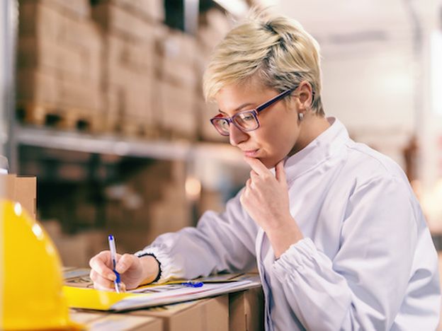 Young blonde caucasian woman in white uniform and with eyeglasses filling out paperwork while standing in warehouse