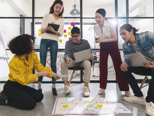 Young creative asian group meeting and looking at project plan lay out on floor discuss or brainstorm business strategy with post note workshop for startup team in small office happy workplace