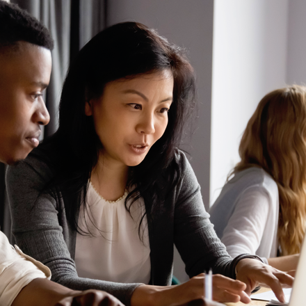Two people looking at a laptop as two other people in the background talk