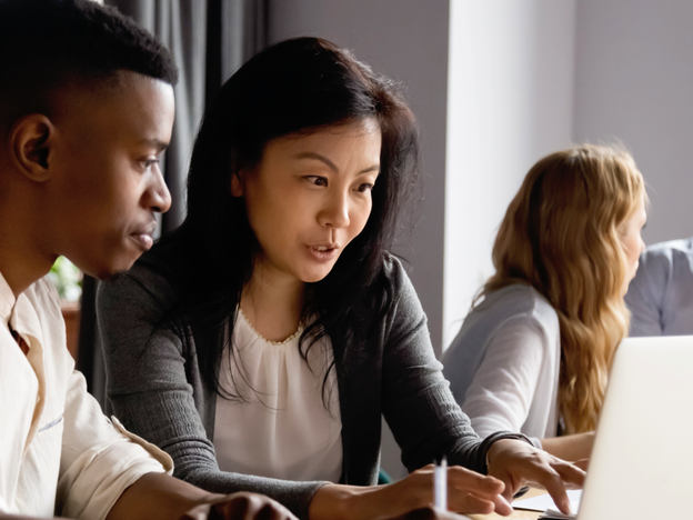 Two people looking at a laptop as two other people in the background talk