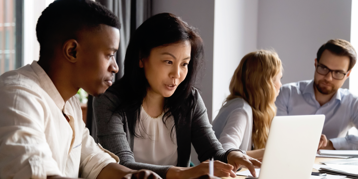 Two people looking at a laptop as two other people in the background talk